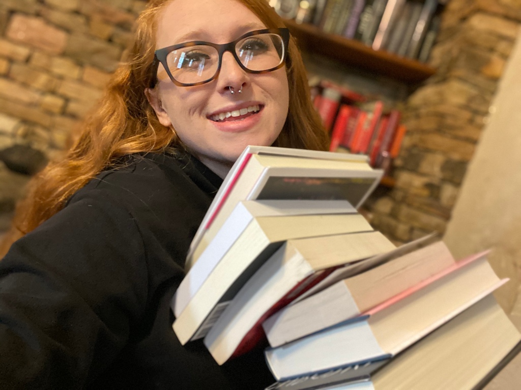 A girl smiling holding a large stack of books up to her chin.  The girl has red hair and black glasses  